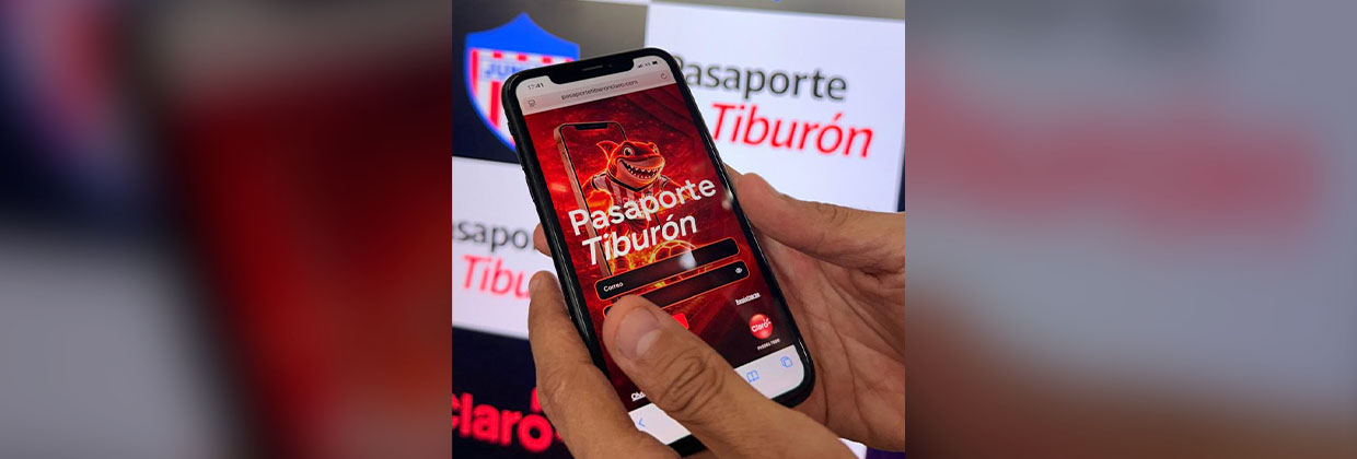 Usuario accede a Pasaporte Tiburón desde su celular junto al escudo del Junior FC Usuario accede a Pasaporte Tiburón desde su celular junto al escudo del Junior FC